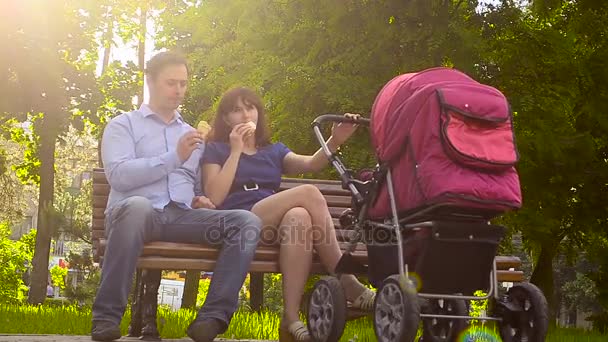 Vader en moeder eten ijs, lopen met baby wandelwagen in park, vakantie met het gezin op de dag af in de zomer — Stockvideo