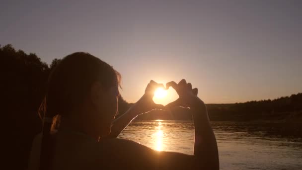 美丽的女孩在河显示手与心脏形状在日落, 反对水的背景 — 图库视频影像