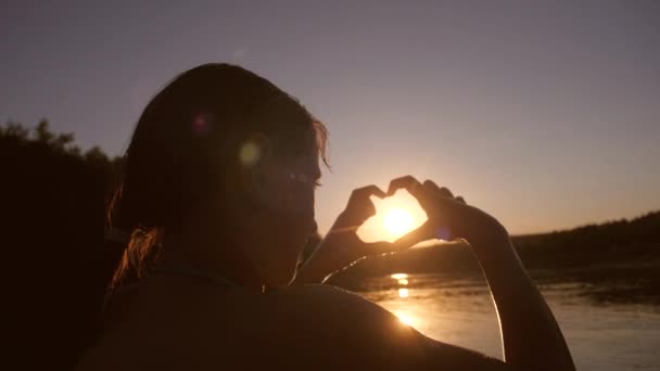 Jong meisje in rivier toont handen met hart vorm bij zonsondergang op de achtergrond van de baai — Stockvideo