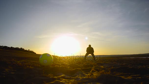 Вратарь ловит мяч на закате, пинает футбольный мяч, силуэты мужчин, играющих в футбол на пляже — стоковое видео