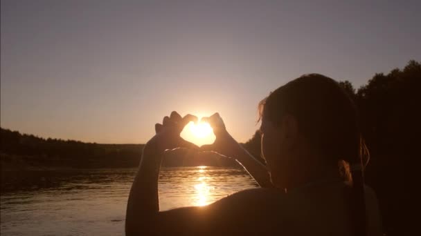 Mooi meisje in rivier toont handen met hart vorm bij zonsondergang, tegen de achtergrond van de water — Stockvideo