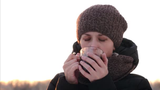 Όμορφο κορίτσι με ευχαρίστηση πίνει ένα ζεστό ρόφημα σε ένα χειμερινό πάρκο και χαμόγελα. — Αρχείο Βίντεο