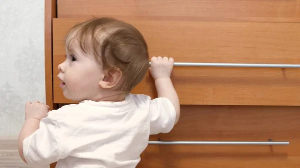 Criança pequena, aprende a andar, segurando o punho do armário, na sala de crianças . — Fotografia de Stock