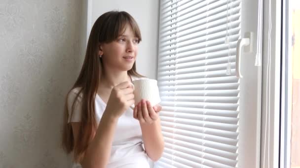 Mujer joven disfrutando de su café o té de la mañana, de pie junto a la ventana y sonriendo. Hermosa chica romántica irreconocible beber bebida caliente en casa acogedora . — Vídeos de Stock