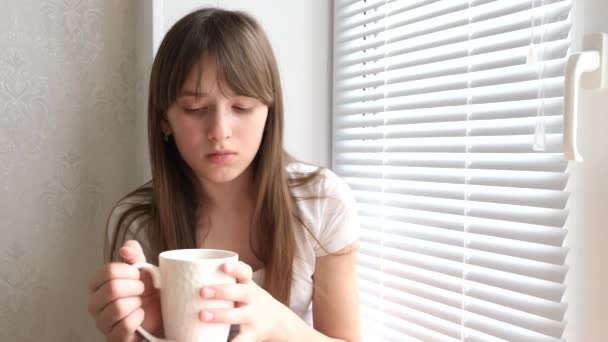 Trauriges Mädchen mit Tasse Kaffee auf Fensterbank am Fenster sitzend. — Stockvideo