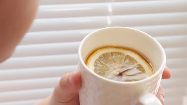 Chica fría bebe té caliente de oro con limón de pie por la ventana . — Vídeo de stock