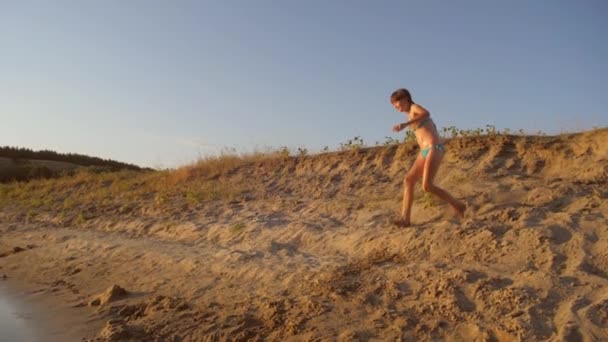 Menina salta da montanha de areia para o rio e banha-se contra o belo pôr do sol no brilho. Movimento lento — Vídeo de Stock