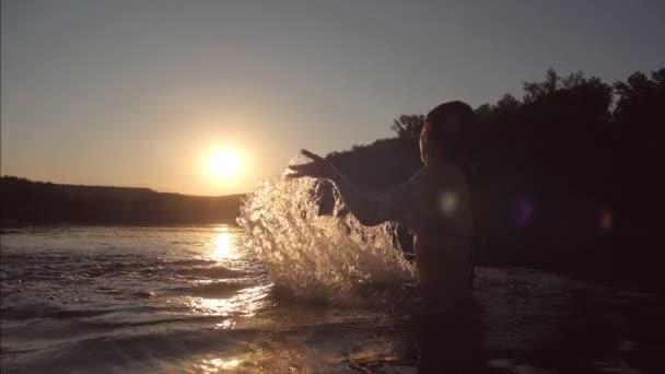 A menina banha-se à noite no rio em frente do pôr-do-sol e espirra a água com uma risada. Movimento lento — Vídeo de Stock