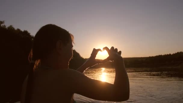 Chica feliz se baña en la noche en el río frente al sol poniente y muestra las manos del corazón . — Vídeos de Stock