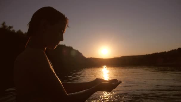 Beautiful girl is played with water standing by river opposite setting sun in the sky smiling — Stock Video