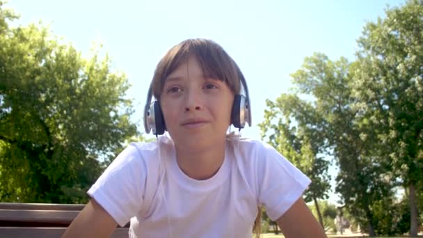 Menina bonita fala através de um microfone fones de ouvido sentado em um banco em um parque . — Vídeo de Stock
