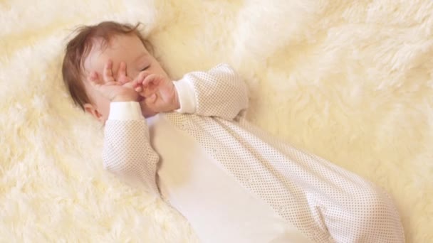 Baby rubs his eyes, lying on his back, on white blanket. — Stock Video