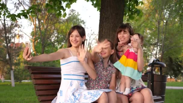 Jong gezin zitten op bankje in park salphy op telefoon glimlachen en zwaaien hun handen doen. — Stockvideo