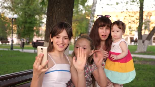 Joven familia sentada en el banco en el parque haciendo salphy en el teléfono sonriendo y agitando las manos . — Vídeos de Stock