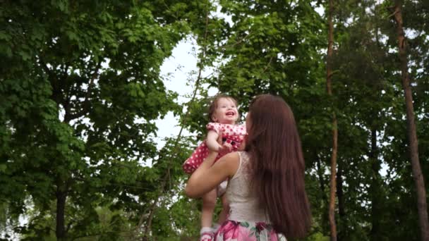 Mädchen lacht in den Armen ihrer Mutter, wenn sie in der Luft fliegt. Mutter hält fröhliches Baby auf dem Arm und geht im grünen Park spazieren. Mütterliches Glück. Zeitlupe. — Stockvideo