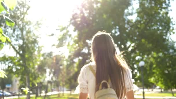 Girl with a backpack goes to park in headphones and listens to music and smiles, teenager happily waves his hand at camera lens. come after me, girl travels around city. Slow motion. — Stock Video