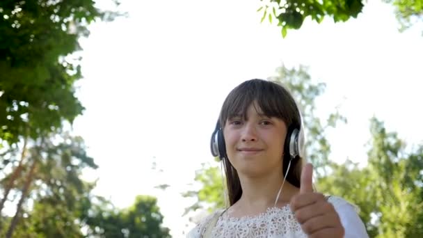 Girl with a backpack goes to park in headphones and listens to music and smiles, teenager happily waves his hand at camera lens. come after me, girl travels around city. Slow motion. — Stock Video