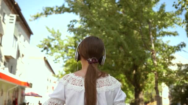 バックパックを持つ女の子は、ヘッドフォンで駐車し、音楽や笑顔を聞いて、ティーンエイジャーは楽しそうにカメラのレンズで手を振ります。私の後に来て、女の子は街を旅行します。スローモーション. — ストック動画