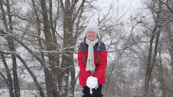 幸せな子供の女の子は森の中で手で雪を投げる。雪が降り、太陽の下で輝きます。子供はクリスマス休暇のために公園で冬に遊ぶ. — ストック動画