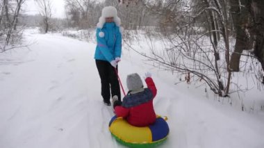 Mutlu anne, beyaz karlı bir yolda kızakla bir çocuğa biniyor. Noel tatilleri. Yetişkinler ve çocuklar için eğlenceli bir oyun. Mutlu aile kavramı. Bir genç Tubing 'e biniyor.