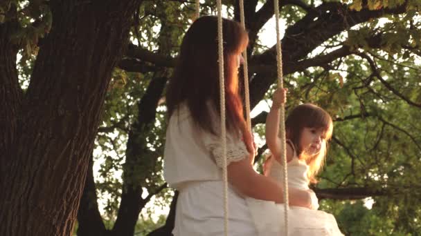 A mãe balança a filha num baloiço debaixo de uma árvore ao sol. close-up. passeio de mãe e bebê em um balanço de corda em um ramo de carvalho na floresta. Ri menina, alegra-se. Diversão familiar no parque, na natureza. dia quente de verão . — Vídeo de Stock