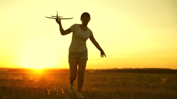 在夕阳西下，快乐的女孩带着一架玩具飞机在田野里奔跑。孩子们玩玩具飞机。青少年的梦想是飞行和成为飞行员。这个女孩想成为一名飞行员和宇航员。慢动作 — 图库视频影像