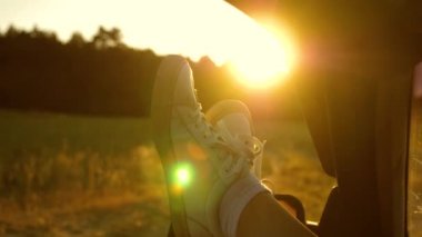 Gün batımında araba camında bir kızın bacakları, güneş ışığı, kır yolunda araba süren bir kızın bacakları. Genç bir kadın, bacaklarını açık bir pencereden dışarı çıkararak arabayla seyahat etmeyi sever. Özgür kadın. Yavaş çekim