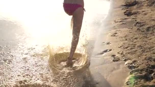 Soleil éclatant. éclaboussures d'eau volent dans différentes directions. Au ralenti. fille court pieds nus le long de la rive et jouer dans l'eau sur la plage au coucher du soleil. adolescent fille avoir amusant en été sur la plage . — Video