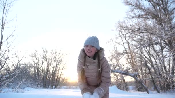 Noel tatili için çocuk parkta oynuyor. Kar yağıyor. Çocuk Noel tatili için parkta oynuyor. Mutlu kız gün batımında kar taneleri fırlatıyor ve kış parkında gülümsüyor. Yavaş — Stok video