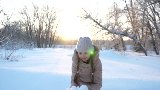 Noel tatili için çocuk parkta oynuyor. Kar yağıyor. Çocuk Noel tatili için parkta oynuyor. Mutlu kız gün batımında kar taneleri fırlatıyor ve kış parkında gülümsüyor. Yavaş — Stok video