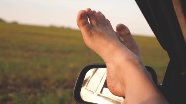 車の窓に女の子の裸の足、太陽のまぶしさ、田舎道で車に乗って。女性は車で旅行する。若い女性は車で旅行するのが好きで、開いた窓から足を出すのが好きです。. — ストック動画