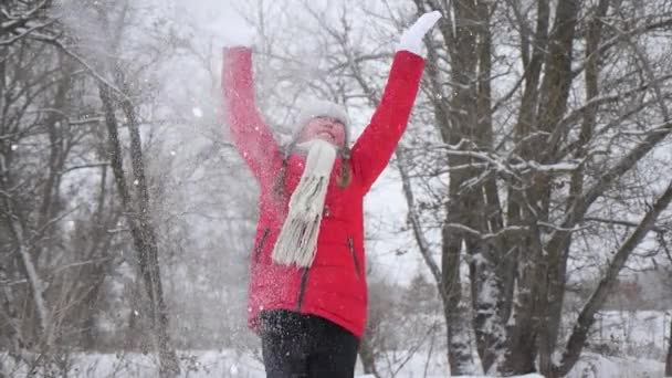 幸せな子供の女の子は森の中で手で雪を投げる。雪が降り、太陽の下で輝きます。子供はクリスマス休暇のために公園で冬に遊ぶ. — ストック動画