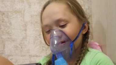 Hastanede yüzünde solunum maskesiyle tedavi edilen küçük bir kız. Tabletli çocuk hasta ve astım spreyinden nefes alıyor. Yakın plan. Yürümeye başlayan çocuk, solunum buharı soluyarak grip tedavisi görüyor..