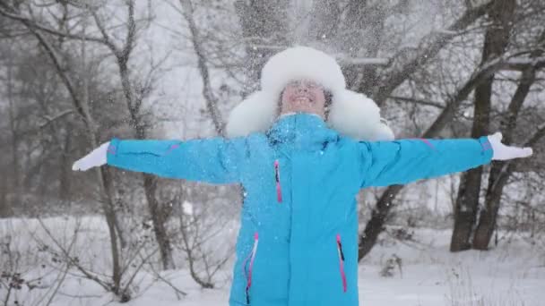 幸せな女性は森の中で手で雪を投げる。雪が降り、太陽の下で輝きます。クリスマス休暇のために公園で冬に遊ぶ女の子。スローモーション — ストック動画