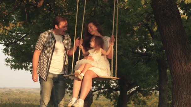 Mam schudt haar dochter op schommel onder een boom in de zon. Papa lacht en verheugt zich. Vader schudt moeder en kind op een touw zwaai op een eik tak in het bos. Familie plezier in het park, in de natuur. — Stockvideo