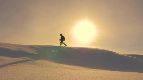 Fotoğrafçı kışın sarı günbatımında karlı tepeye çıkar. Kameralı ve tripodlu dağcı, kar üzerinde yürüyor.. — Stok fotoğraf