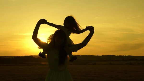 Mamma e bambino felici attraversano il campo al tramonto e ridono. Una bambina si siede sulle spalle della madre e cammina insieme al parco al sole e sorride. Concetto di vita familiare. Rallentatore — Foto Stock