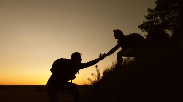 女性旅行者は丘の上に登るのを助ける男性旅行者の手を握っています。観光客の手を握って、日没で山に登る。ビジネスパートナーのチームワークです休暇中の幸せな家族. — ストック写真