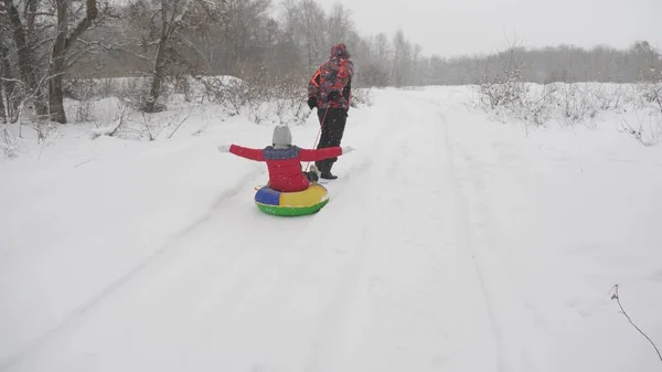 Glad pappa släpar ett barn på en snöig väg. Julhelger. Pappa leker med sin dotter i en vinterpark. Begreppet lycklig familj. Tonårsturer i Tubing — Stockfoto