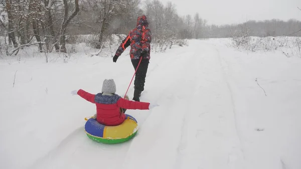 Joyeux papa traîne un enfant sur une route enneigée. Vacances de Noël. père joue avec sa fille dans un parc d'hiver. Le concept d'une famille heureuse. adolescents promenades dans Tubing — Photo