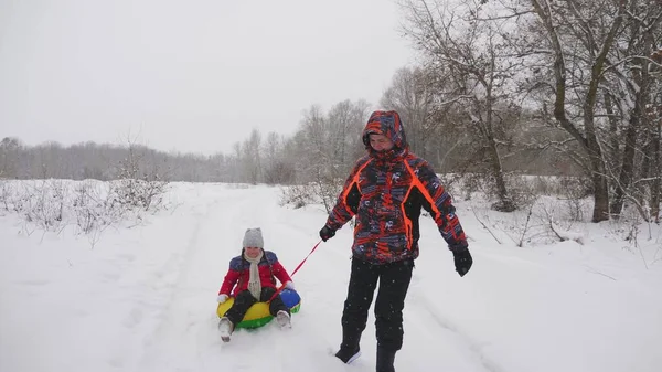 Der glückliche Vater fährt ein Kind auf einem Schlitten auf einer weißen, schneebedeckten Straße. Weihnachtsferien. ein lustiges Spiel für Erwachsene und Kinder. das Konzept einer glücklichen Familie. Teenager fährt in Schläuchen — Stockfoto