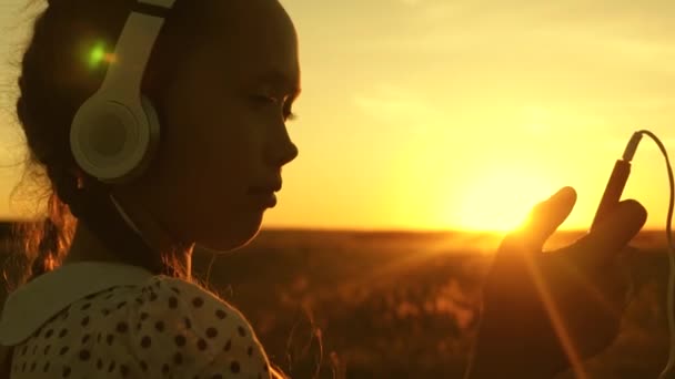 Barn reser och lyssnar på musik i varm sol. Tonårstjejen lyssnar på musik och tittar på solnedgången. Lycklig flicka dansar i hörlurar i strålarna av en vacker soluppgång i parken. — Stockvideo