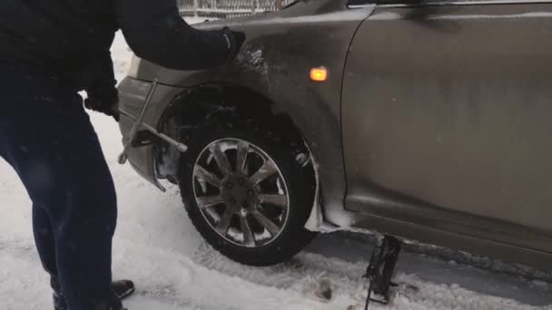 Mężczyzna podnosi jack samochodu na drodze. człowiek jest zmiana koła samochodu. Wymiana koła w zimie na drodze w blizzard i śniegu. Podział samochodów, wymiana koła. — Wideo stockowe
