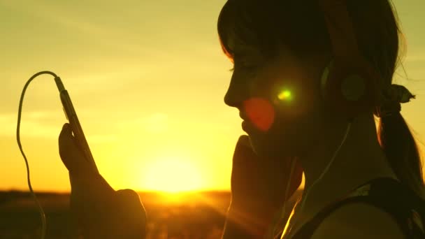 Lycklig flicka dansar i hörlurar i strålarna av en vacker soluppgång i parken. flicka reser och lyssnar på musik i den varma solen. tonåring flicka lyssnar på musik och tittar på solnedgången. — Stockvideo