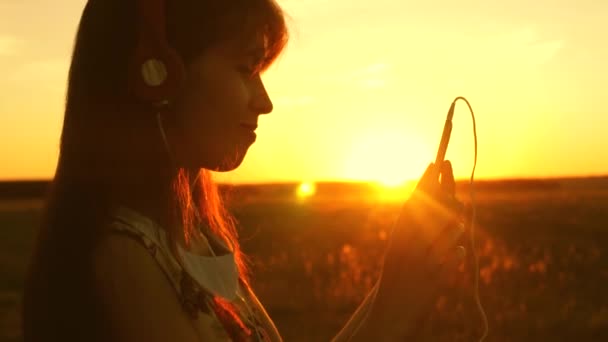 Belle femme voyage en écoutant de la musique et en dansant dans les rayons d'un beau coucher de soleil. jeune fille dans les écouteurs et avec un smartphone touche doigt au capteur de la tablette sélectionne des chansons en ligne. Doucement. — Video