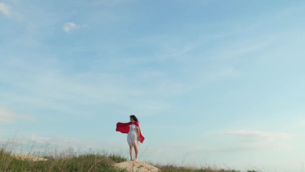 Jonge vrouw die dolt en grappen maakt in een rode cape. Meisje droomt ervan om een superheld te worden. Sexy superheld meisje staat op het veld in een rode mantel, mantel fladdert in de wind. — Stockvideo
