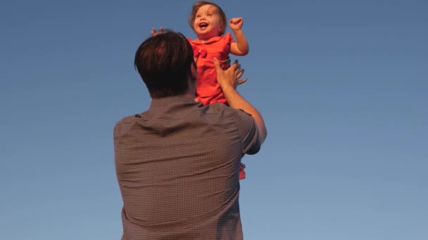 Vader geeft zijn kind over in de blauwe lucht Papa speelt met een dochtertje, de baby lacht en verheugt zich. Langzame beweging. gelukkig gezin spelen in de avond tegen de hemel. — Stockvideo
