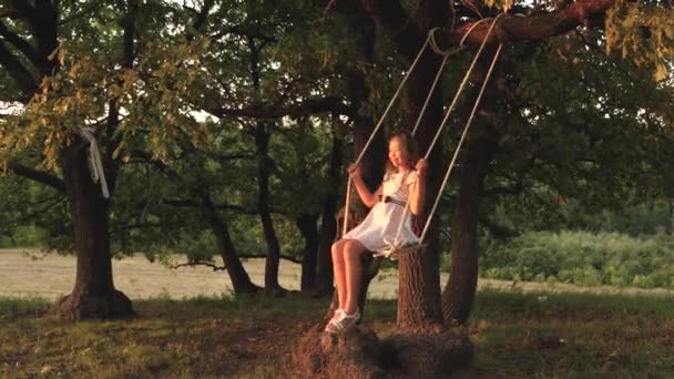 Giovane ragazza che oscilla su un'altalena sotto un albero al sole, giocando con i bambini. primo piano. Divertimento in famiglia nella natura. bambino cavalca un'altalena corda su un ramo di quercia nel parco il tramonto. ragazza ride, gioisce . — Video Stock