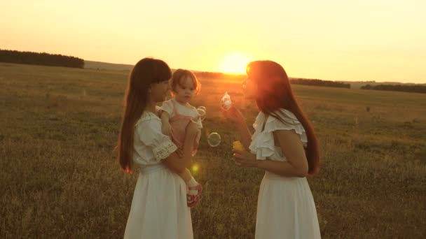 Las hijas se alegran y sonríen, las burbujas vuelan en el parque al atardecer. En cámara lenta. Feliz madre jugando con los niños soplando burbujas de jabón. concepto de una familia feliz. niño, hermana y madre juegan al amanecer . — Vídeos de Stock