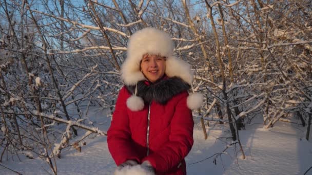 Bella ragazza adolescente che gioca in un parco invernale. ragazza felice getta la neve con le mani al tramonto nella foresta. Cadute di neve e scintille al sole . — Video Stock